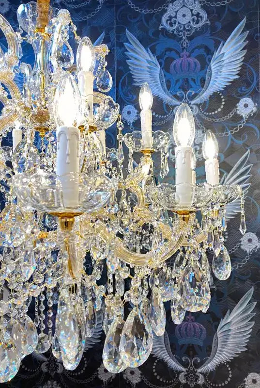 Exklusiver Kronleuchter Munich Gold Ø95cm mit 18 Kristallglas-Armen vor blauem Hintergrund, beleuchtet mit LED-Lampen.
