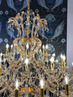 Luxus Kronleuchter Gold Maria Theresia with 42 glass-clad arms, featuring sparkling crystals against a decorative wall background.